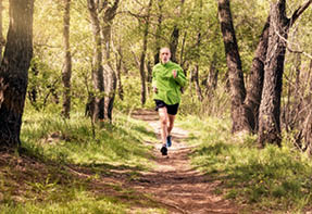 A senior man worn in black and green is running in the forest  during a warm spring day