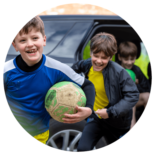 A three-quarter length of a mother opening the car door for her children on the drive. The children are running out of the car wearing football attire.
