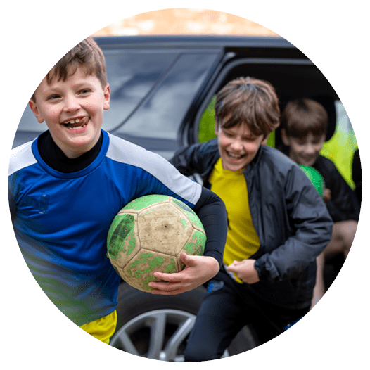 A three-quarter length of a mother opening the car door for her children on the drive. The children are running out of the car wearing football attire.