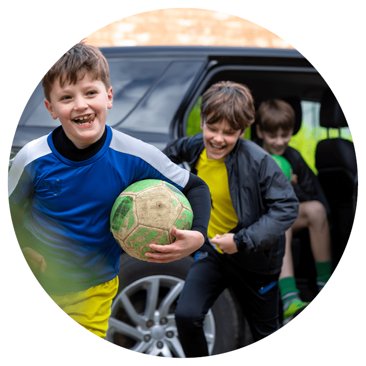 A three-quarter length of a mother opening the car door for her children on the drive. The children are running out of the car wearing football attire.