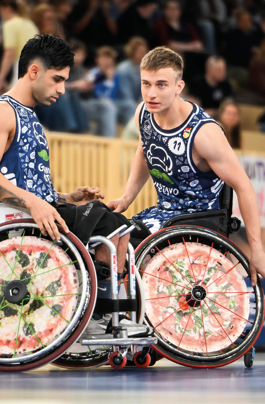 Bilder zweier Profi-Rollstuhlbasketballer w hrend eines Match. Auf den R dern der beiden Rollst hle ist eine gro e Pizza abgeblidet als Werbeanzeige f r einen Lieferservice