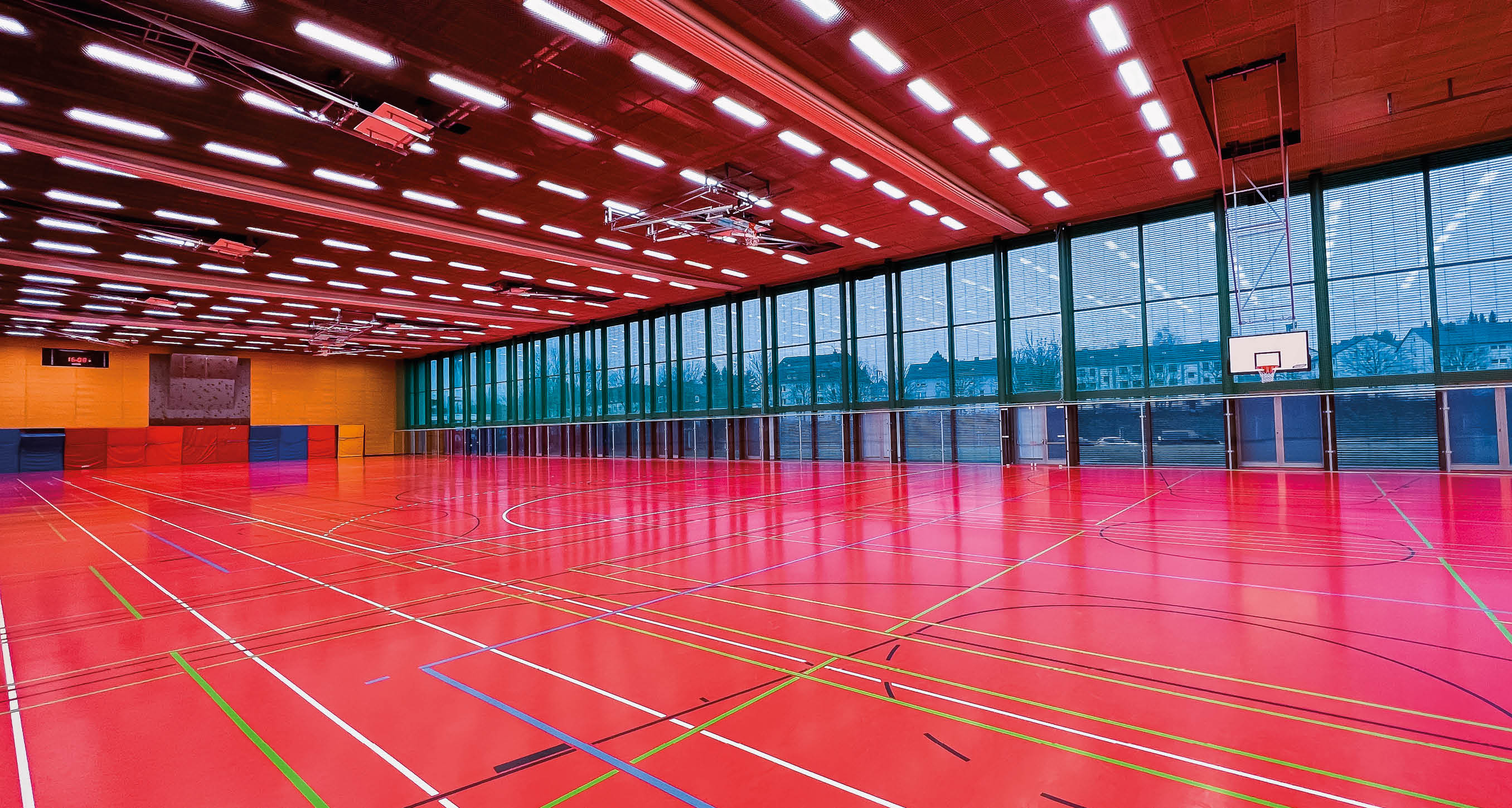 Bild einer leeren Sporthalle. Zu sehen ist der rote Hallenboden mit den markiereungen f r verschiedene Spielfeder. Eine  braune Holzwand im Hintergrund und im rechten Bereich eine Fensterfront.