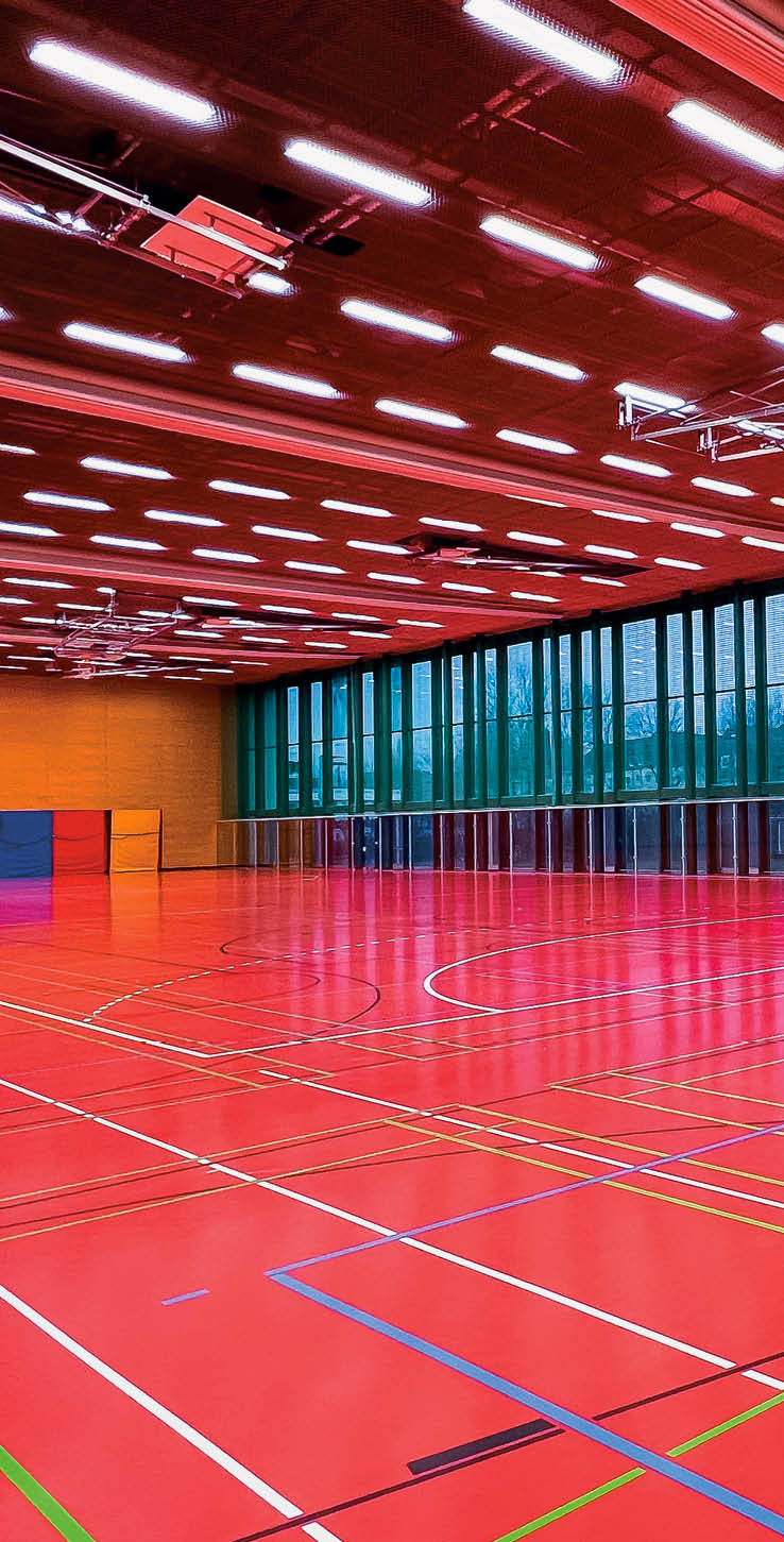 Bild einer leeren Sporthalle. Zu sehen ist der rote Hallenboden mit den markiereungen f r verschiedene Spielfeder. Eine  braune Holzwand im Hintergrund und im rechten Bereich eine Fensterfront.