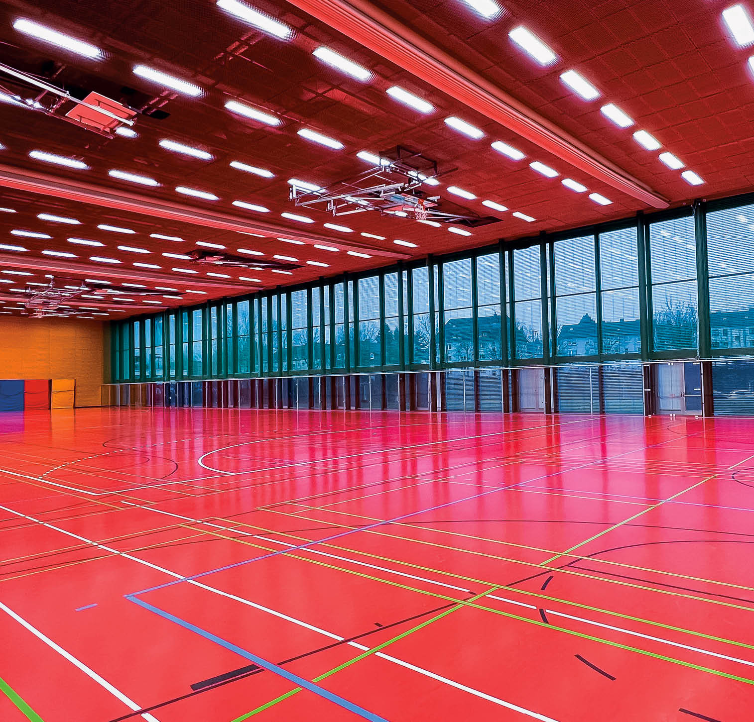 Bild einer leeren Sporthalle. Zu sehen ist der rote Hallenboden mit den markiereungen f r verschiedene Spielfeder. Eine  braune Holzwand im Hintergrund und im rechten Bereich eine Fensterfront.