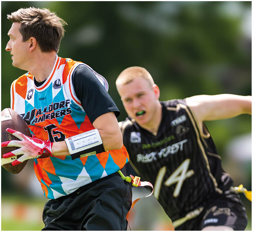 Zwei Personen spielen Flag Football im Freien, eine tr gt ein buntes Trikot und h lt den Ball, w hrend die andere in einem schwarzen Trikot versucht, die Flagge zu greifen. Beschreibung von KI generiert