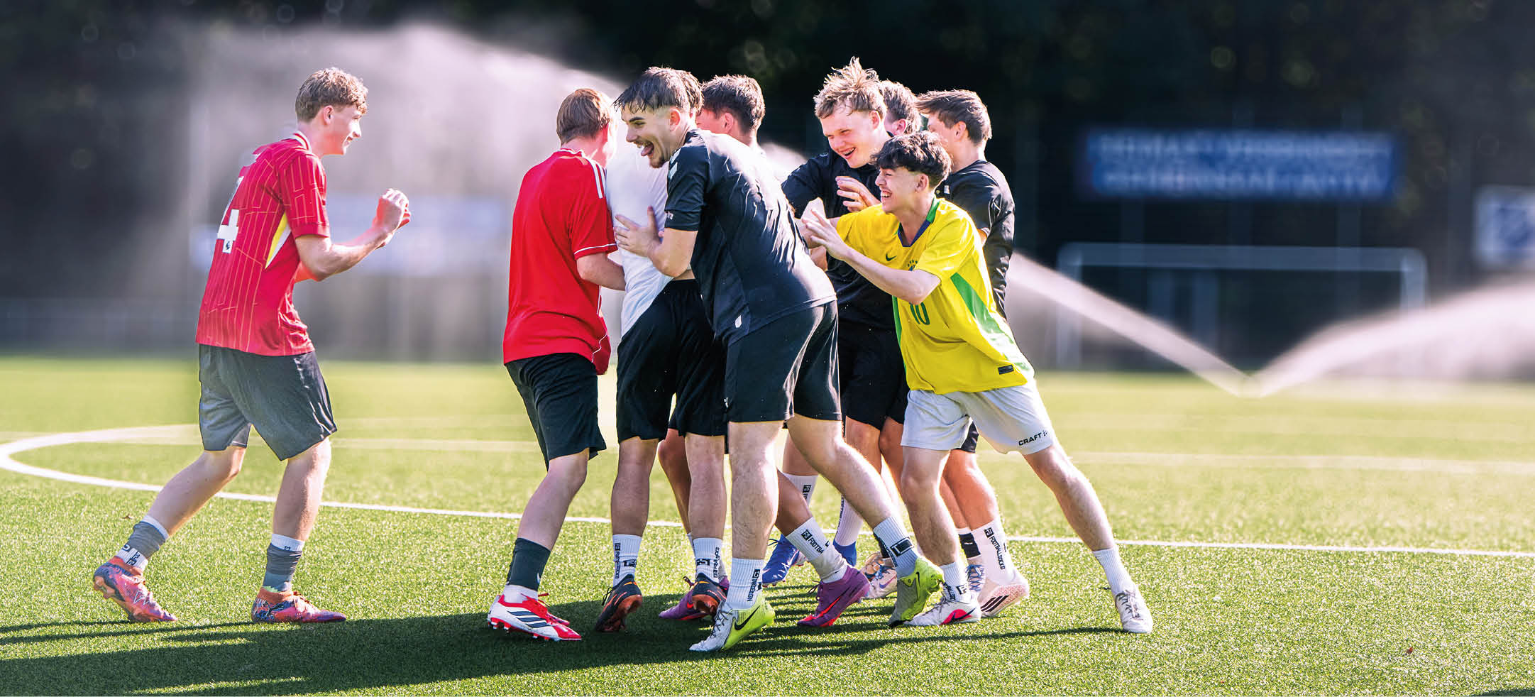 Eine Gruppe von Erwachsenen in Sportkleidung steht lachend und umarmend auf einem Fu ballfeld bei Sonnenschein. Beschreibung von KI generiert