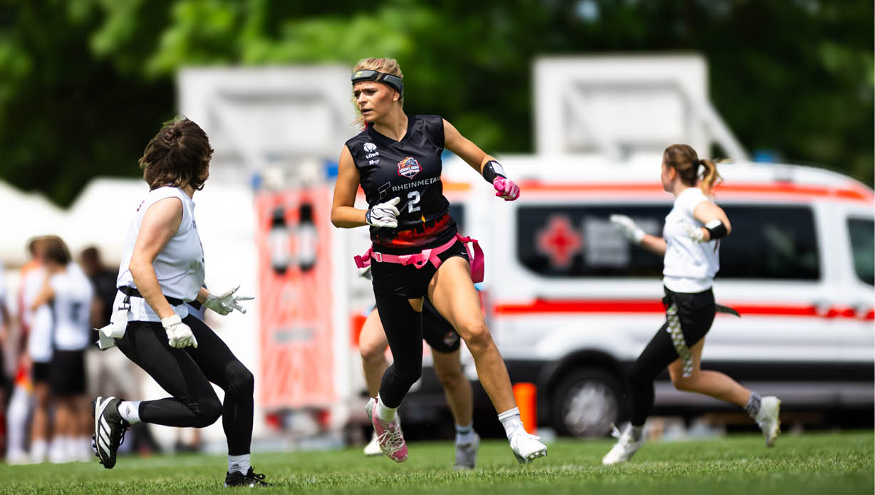 Nika wiesner und weitere Mitspieler spielen bei sonnigem Wetter ein Flag-Football-Spiel auf einem Spielfeld mit einem Krankenwagen im Hintergrund. 