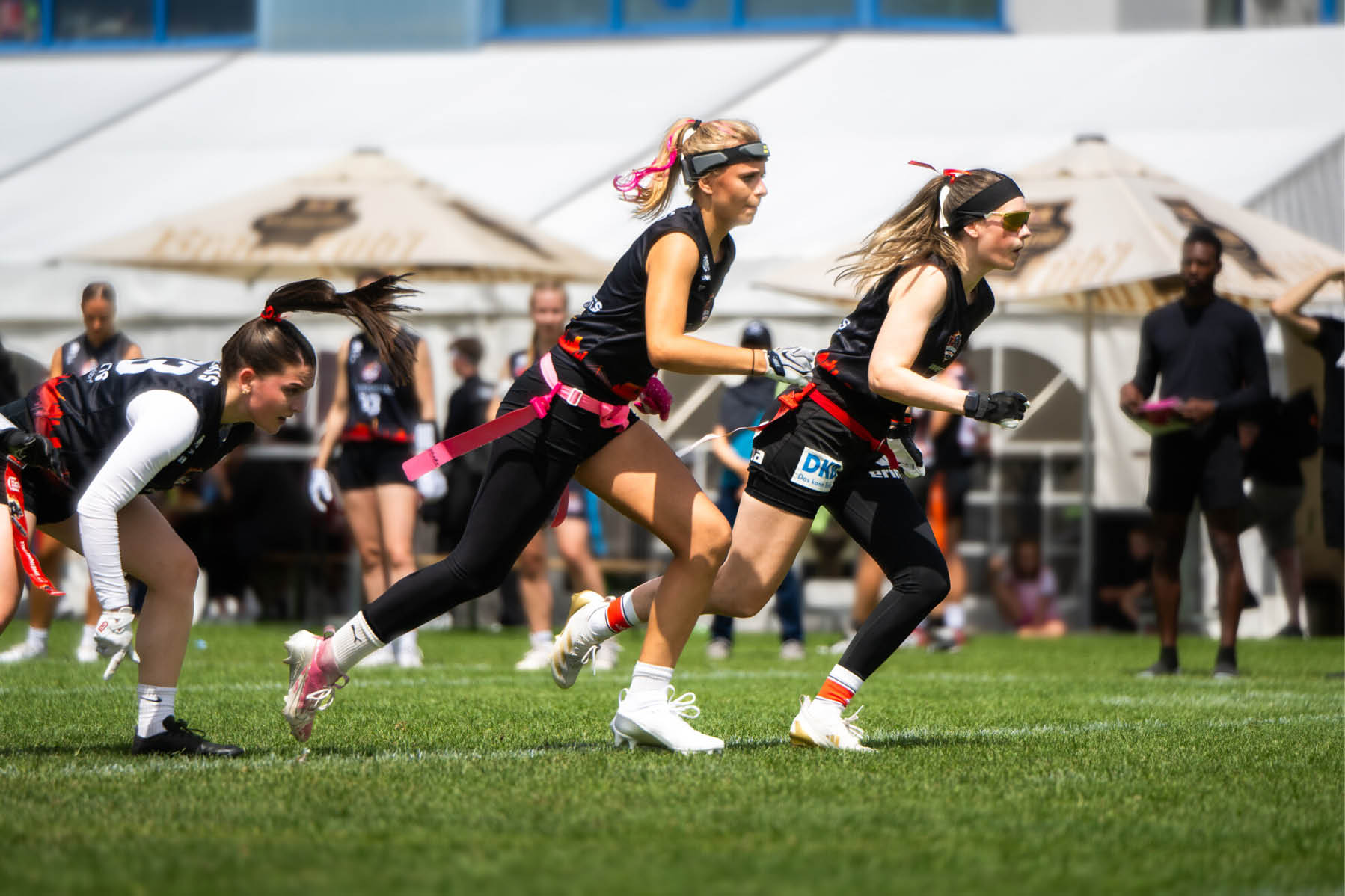 Mehrere Erwachsene in Sportkleidung rennen auf einem Spielfeld, w hrend im Hintergrund Zuschauer unter Sonnenschirmen sitzen. Beschreibung von KI generiert