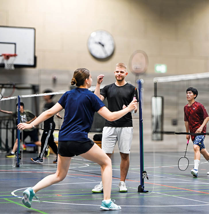Mehrere Personen spielen in einer Halle Badminton, wobei eine Person in blauer Sportkleidung aktiv einen Schlag ausf hrt und eine andere Person in schwarzem Shirt und wei en Shorts zuschaut. Beschreibung von KI generiert