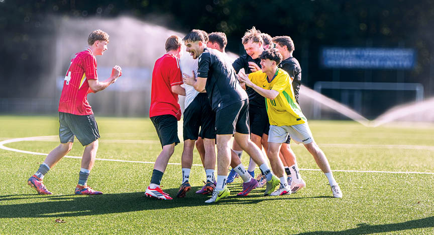 Eine Gruppe von Erwachsenen in Sportkleidung steht lachend und umarmend auf einem Fu ballfeld bei Sonnenschein. Beschreibung von KI generiert