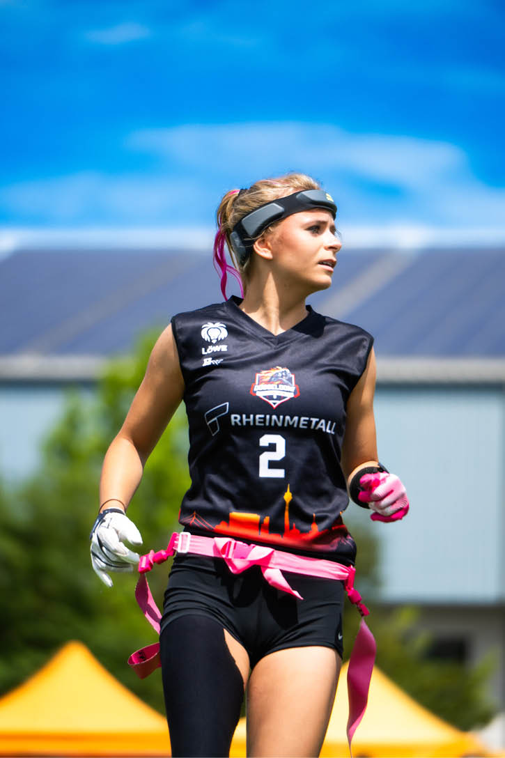 Eine Person tr gt ein schwarzes Sporttrikot mit der Nummer 2 und pinken Flaggenb ndern und steht im Freien vor einem Geb ude mit Solarpaneelen auf dem Dach. Beschreibung von KI generiert