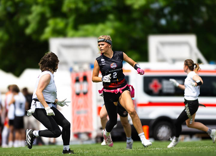 Nika wiesner und weitere Mitspieler spielen bei sonnigem Wetter ein Flag-Football-Spiel auf einem Spielfeld mit einem Krankenwagen im Hintergrund. 