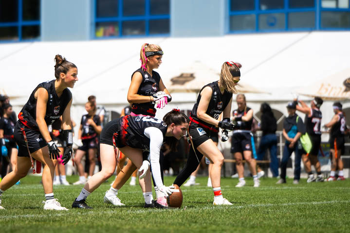 Mehrere Erwachsene in Sportkleidung bereiten sich auf einem Spielfeld auf den Start eines Spiels mit einem Football vor, w hrend im Hintergrund weitere Personen zuschauen. Beschreibung von KI generiert
