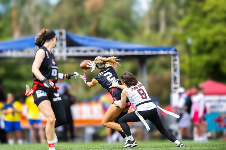 Drei Personen in Sportkleidung spielen American Football auf einem Spielfeld, wobei eine Person den Ball h lt und eine andere versucht, sie zu stoppen. Beschreibung von KI generiert