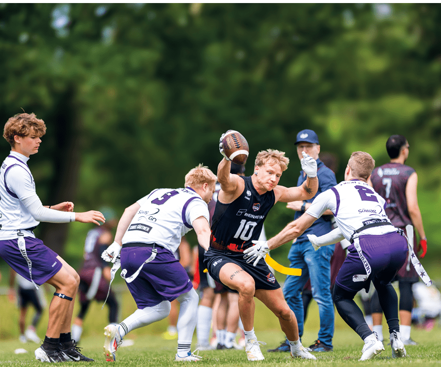 Mehrere Personen in Sportkleidung spielen auf einem Rasenfeld American Football, wobei eine Person den Ball h lt und von anderen umringt wird. Beschreibung von KI generiert