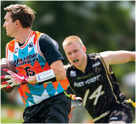Zwei Personen spielen Flag Football im Freien, eine tr gt ein buntes Trikot und h lt den Ball, w hrend die andere in einem schwarzen Trikot versucht, die Flagge zu greifen. Beschreibung von KI generiert