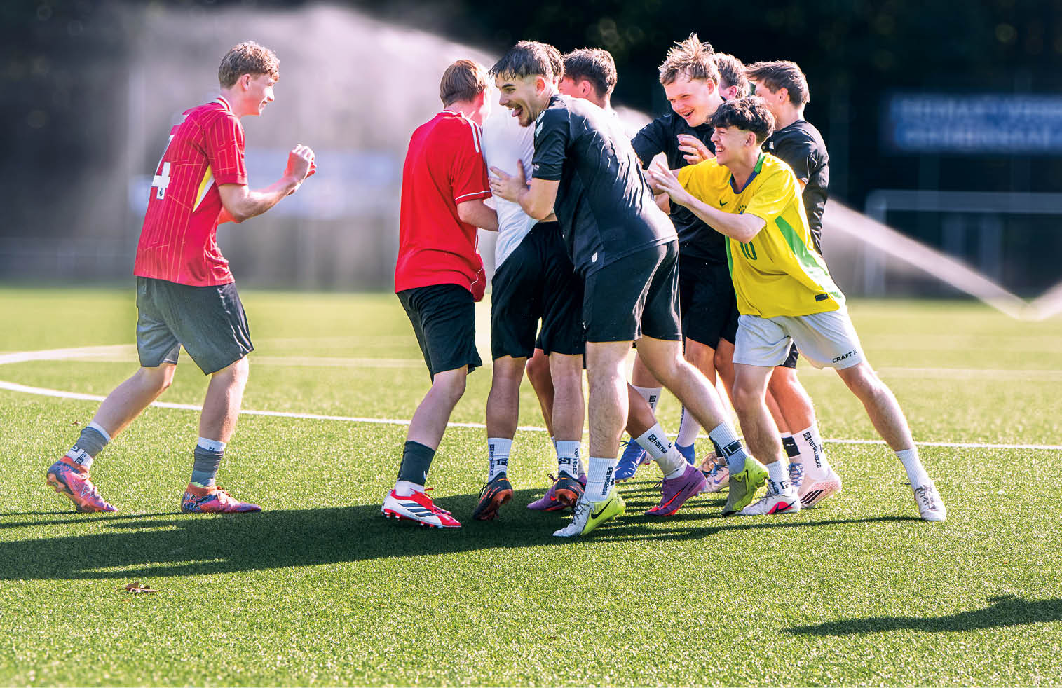 Eine Gruppe von Erwachsenen in Sportkleidung steht lachend und umarmend auf einem Fu ballfeld bei Sonnenschein. Beschreibung von KI generiert