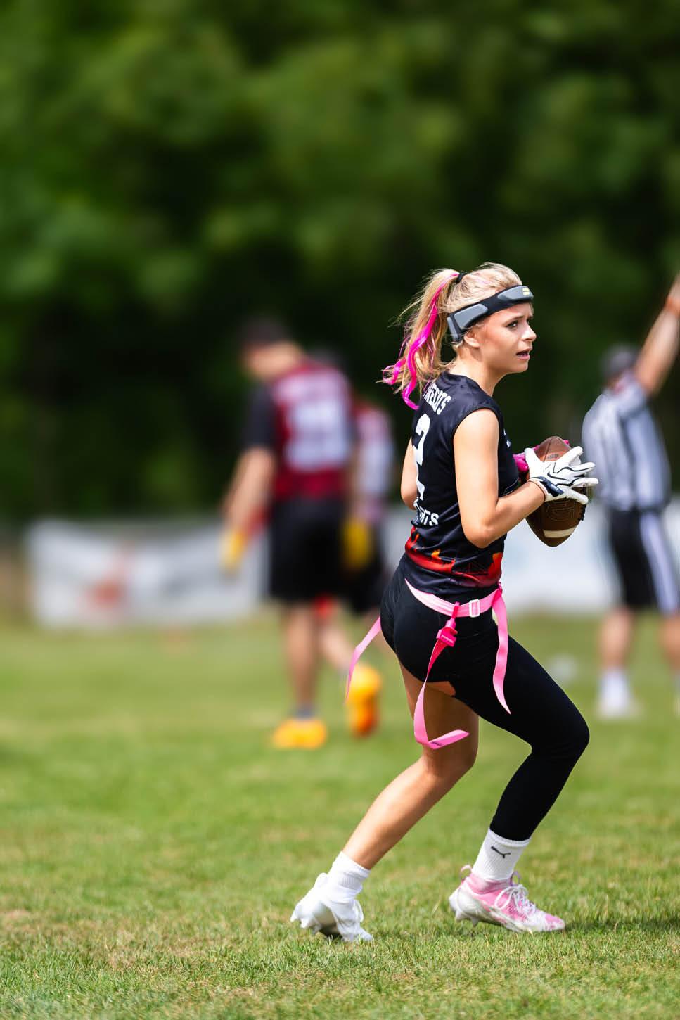 Eine Person mit pinken Haarstr hnen tr gt Sportkleidung und h lt einen Football auf einem Spielfeld w hrend eines Spiels mit weiteren Personen im Hintergrund. Beschreibung von KI generiert