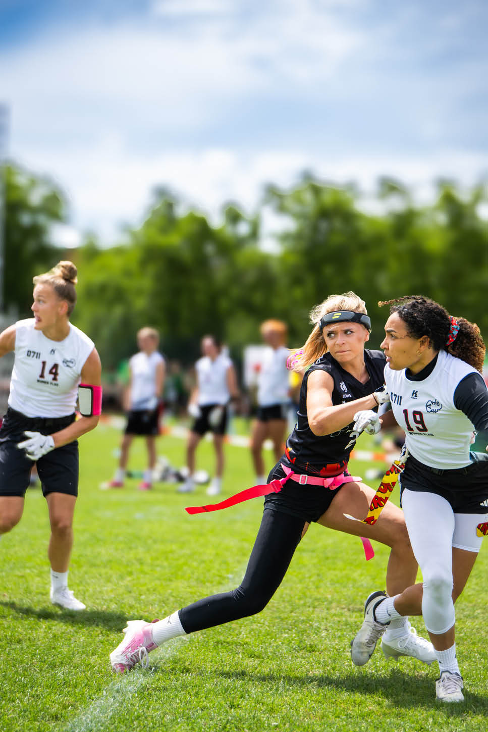 Mehrere Personen spielen auf einem gr nen Spielfeld Flag Football, wobei zwei von ihnen in Aktion sind und versuchen, sich gegenseitig die Flaggen abzunehmen. Beschreibung von KI generiert