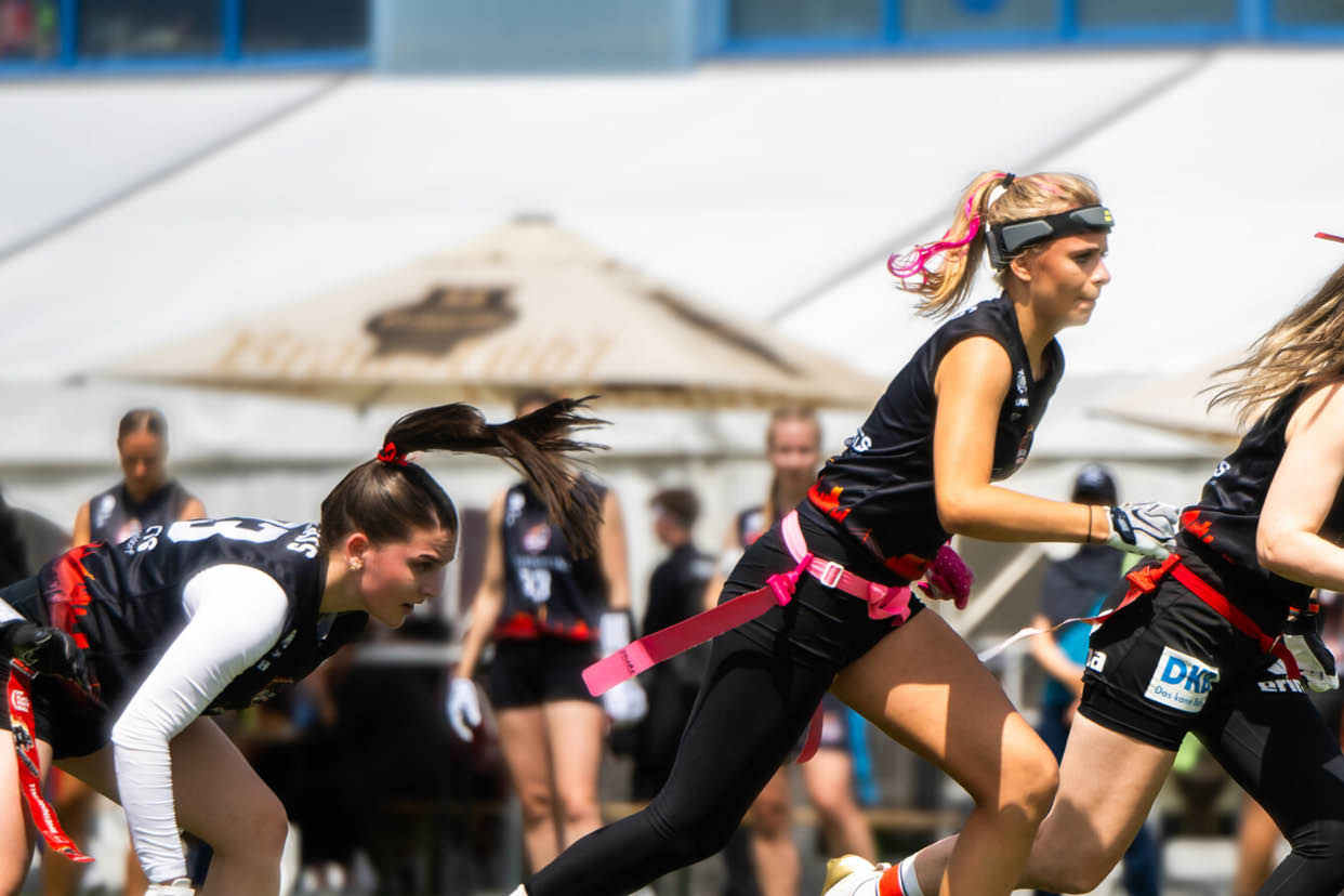 Mehrere Erwachsene in Sportkleidung rennen auf einem Spielfeld, w hrend im Hintergrund Zuschauer unter Sonnenschirmen sitzen. Beschreibung von KI generiert
