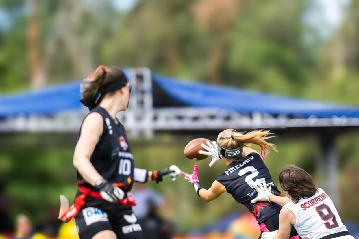 Drei Personen in Sportkleidung spielen American Football auf einem Spielfeld, wobei eine Person den Ball h lt und eine andere versucht, sie zu stoppen. Beschreibung von KI generiert