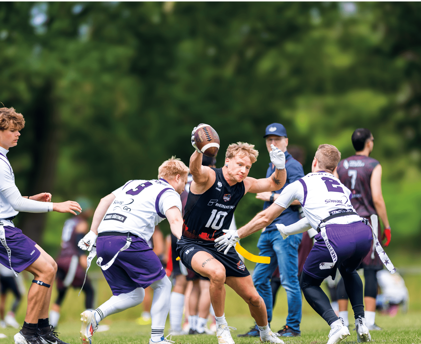 Mehrere Personen in Sportkleidung spielen auf einem Rasenfeld American Football, wobei eine Person den Ball h lt und von anderen umringt wird. Beschreibung von KI generiert