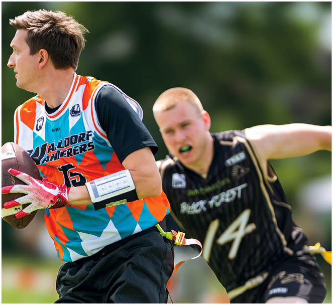 Zwei Personen spielen Flag Football im Freien, eine tr gt ein buntes Trikot und h lt den Ball, w hrend die andere in einem schwarzen Trikot versucht, die Flagge zu greifen. Beschreibung von KI generiert