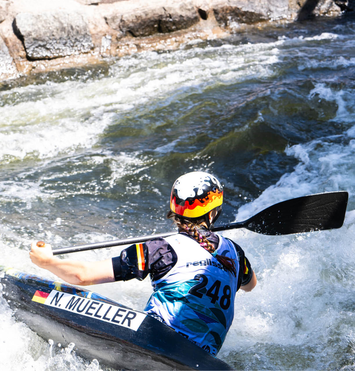 Eine Person mit Helm und geflochtenem Haar paddelt in einem Wildwasserfluss. Beschreibung von KI generiert