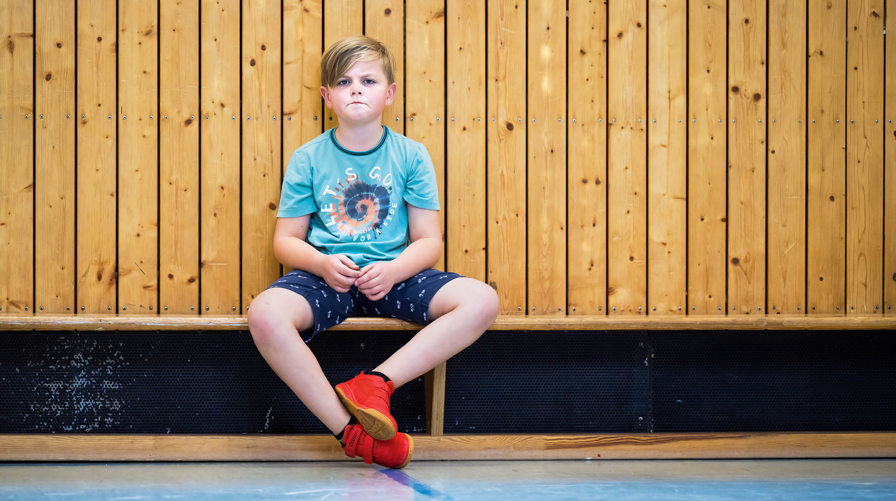 Ein Kind mit kurzem Haar sitzt in einem t rkisfarbenen T-Shirt und dunklen Shorts mit roten Sportschuhen vor einer Holzverkleidung und schaut ernst.