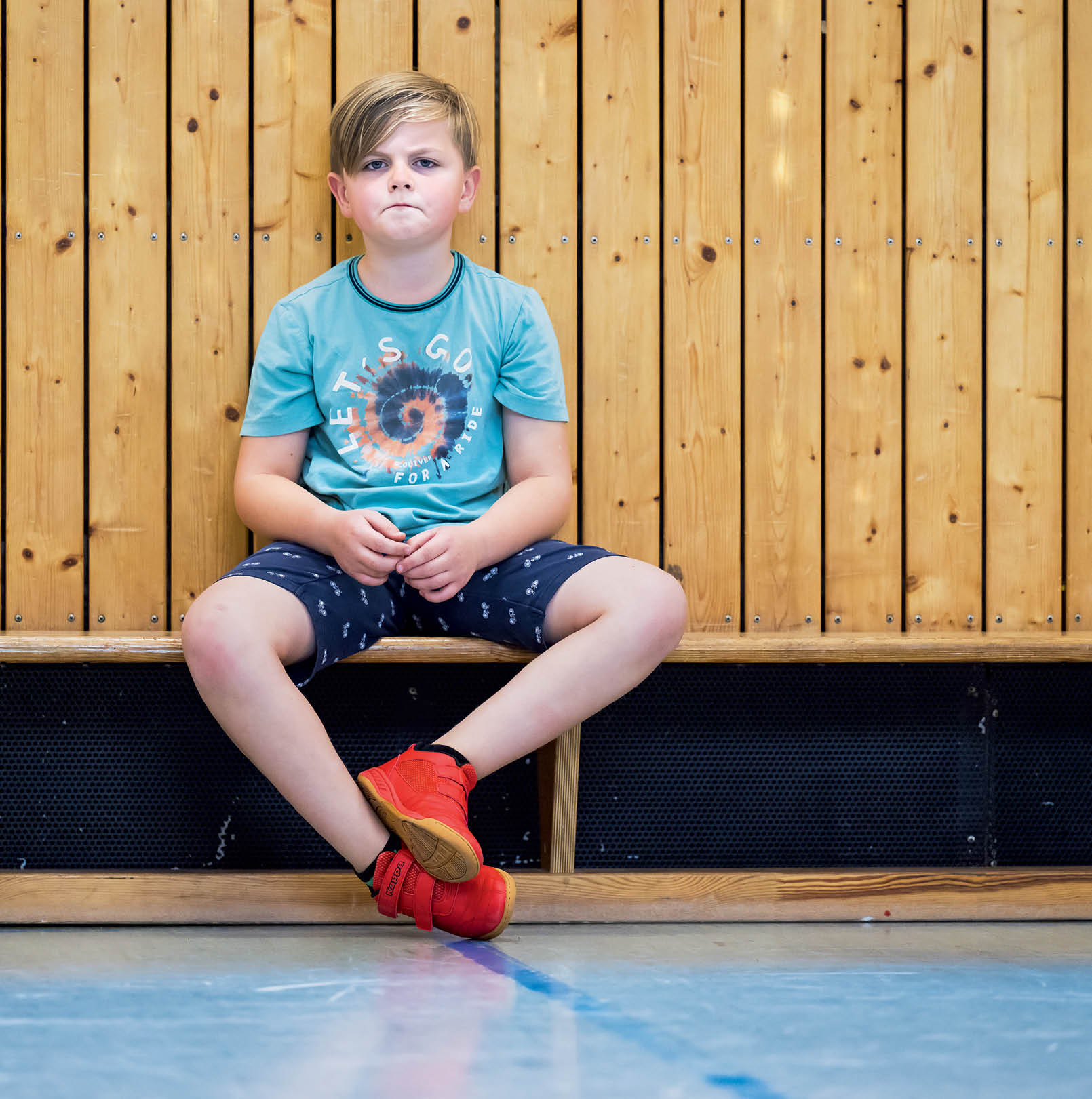 Ein Kind mit kurzem Haar sitzt in einem t rkisfarbenen T-Shirt und dunklen Shorts mit roten Sportschuhen vor einer Holzverkleidung und schaut ernst.