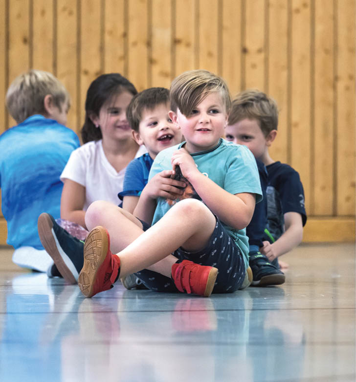 F nf Kinder sitzen in einer Reihe auf dem Boden eines Raums mit Holzwand, wobei das vorderste Kind ein Spielzeug h lt und alle entspannt wirken. Beschreibung von KI generiert