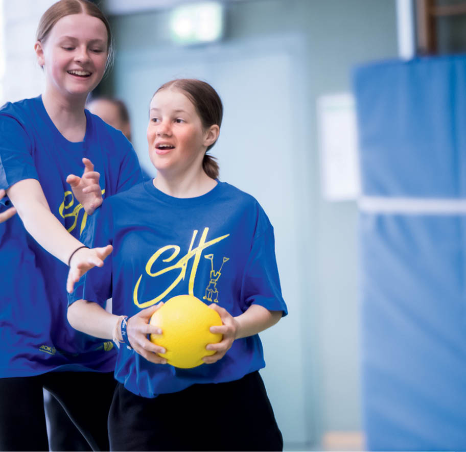 Zwei Personen in blauen T-Shirts spielen gemeinsam mit einem gelben Ball in einer hellen Sporthalle. Beschreibung von KI generiert