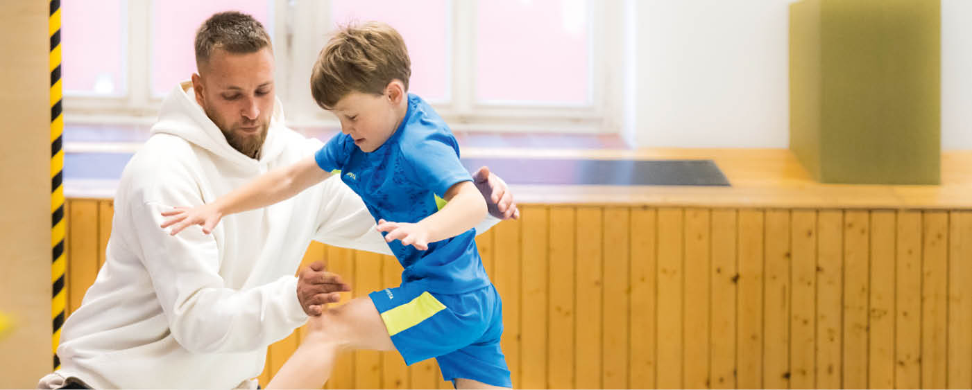 Ein Erwachsener in wei em Kapuzenpullover unterst tzt ein Kind in blauem Sportoutfit beim Balancieren oder Springen in einem hellen Raum mit Holzverkleidung. Beschreibung von KI generiert