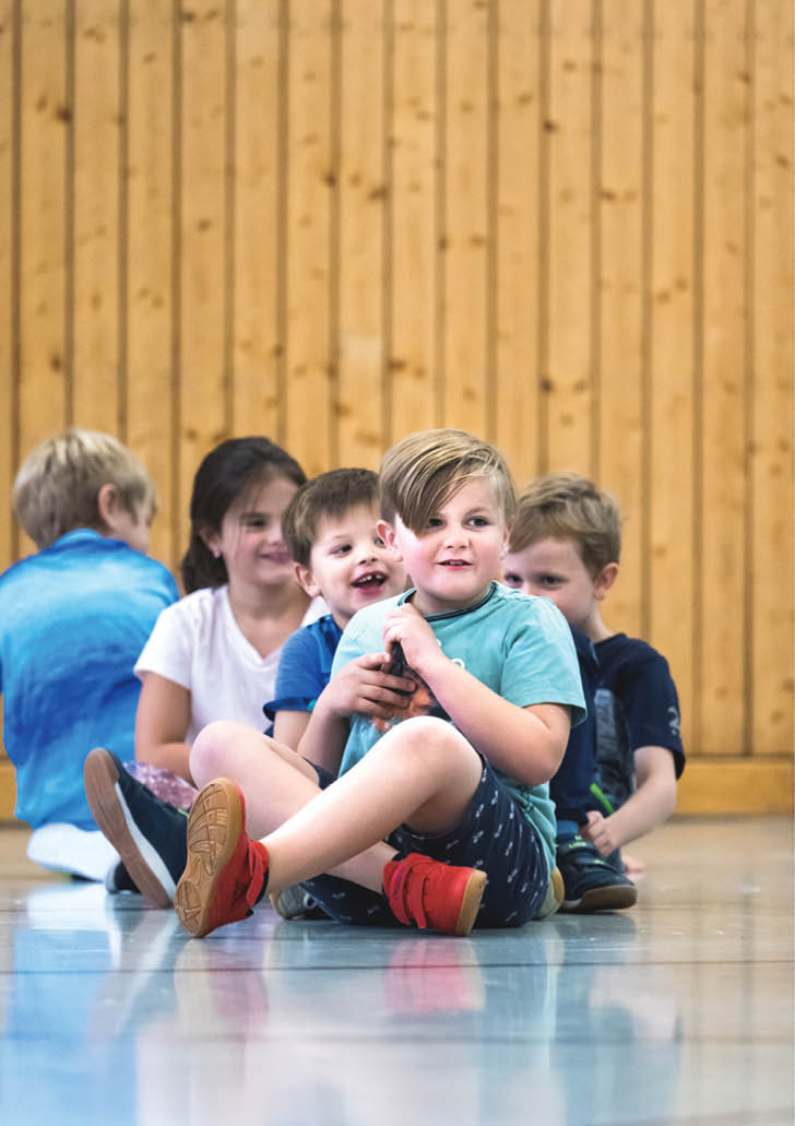 F nf Kinder sitzen in einer Reihe auf dem Boden eines Raums mit Holzwand, wobei das vorderste Kind ein Spielzeug h lt und alle entspannt wirken. Beschreibung von KI generiert