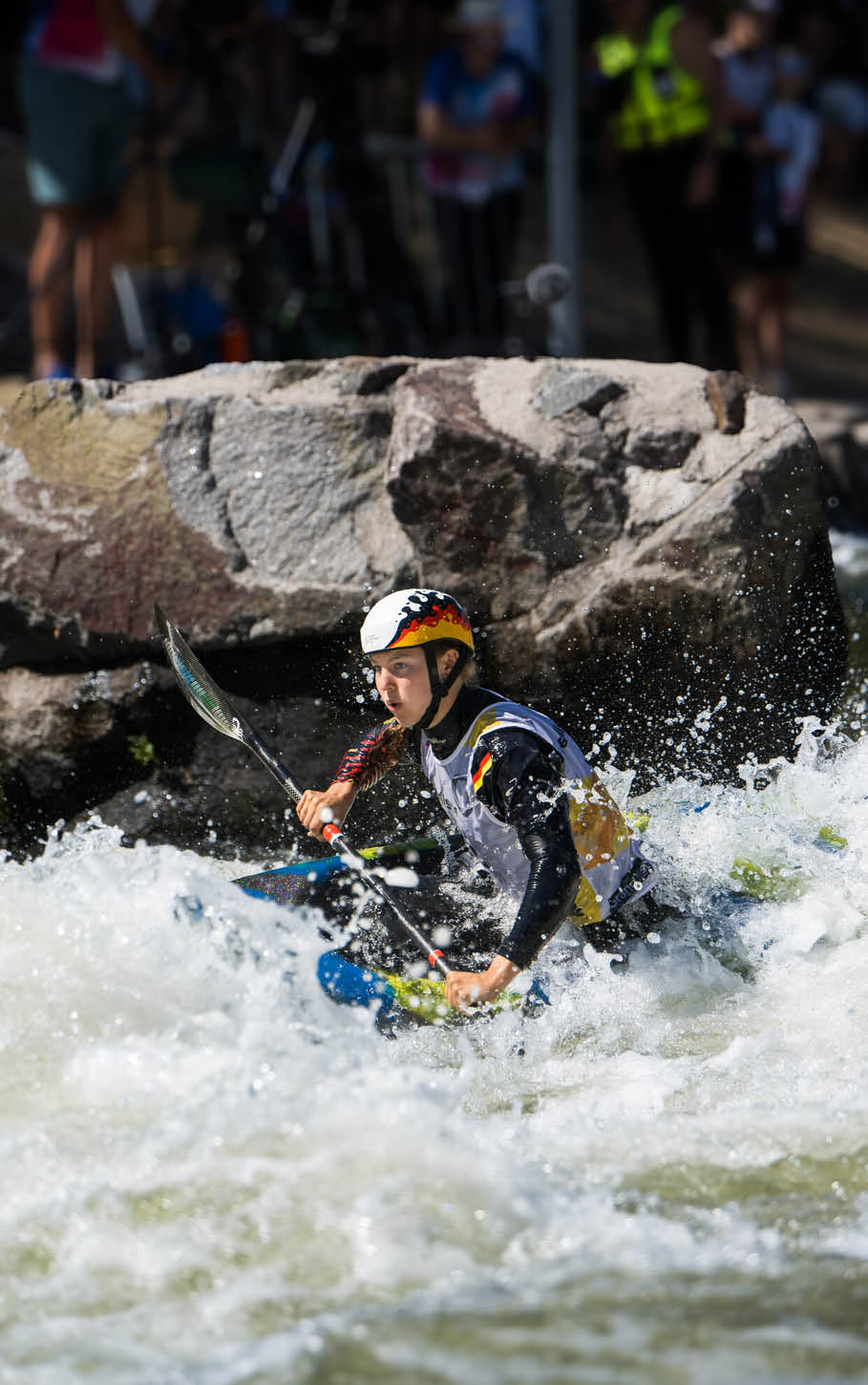 Eine Person mit Helm und Schwimmweste paddelt konzentriert in einem Kajak durch wildes, spritzendes Wasser vor einem gro en Felsen. Beschreibung von KI generiert