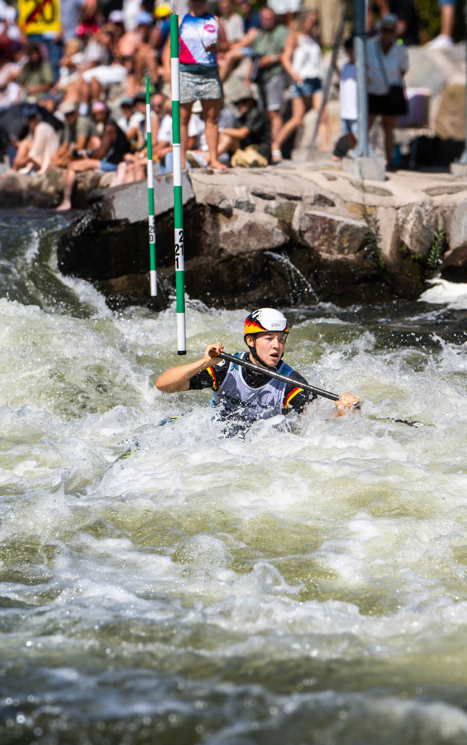 Eine Person tr gt einen Helm und Paddel und f hrt in einem Wildwasserfluss durch ein Tor bei einem Kanuslalom-Wettbewerb vor einer Zuschauergruppe. Beschreibung von KI generiert