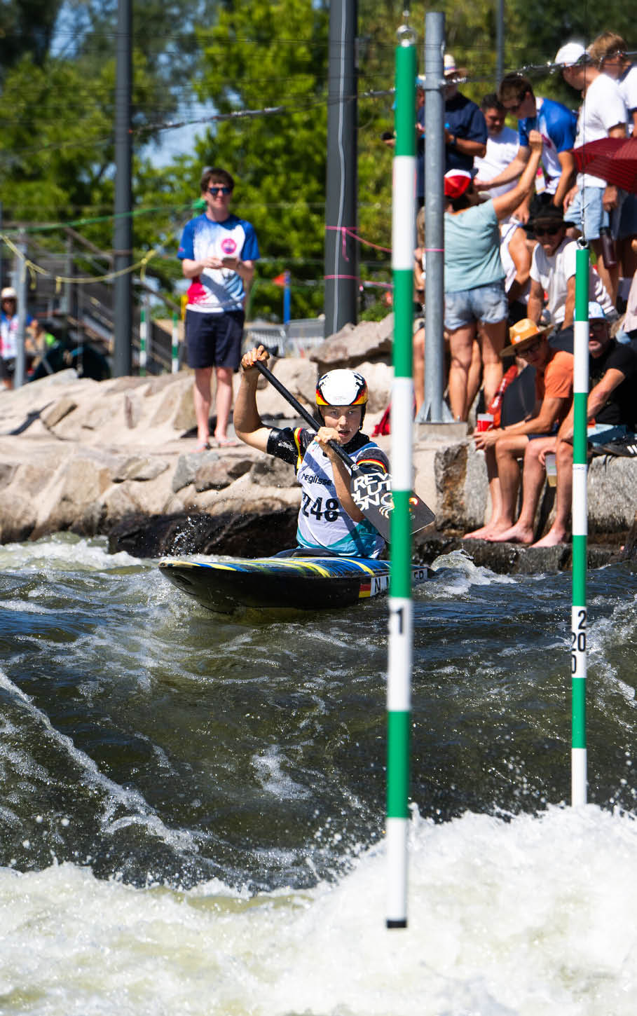 Eine Person mit Helm und Sportbekleidung paddelt in einem Kajak durch Wildwasser, w hrend mehrere Menschen am Ufer zuschauen. Beschreibung von KI generiert