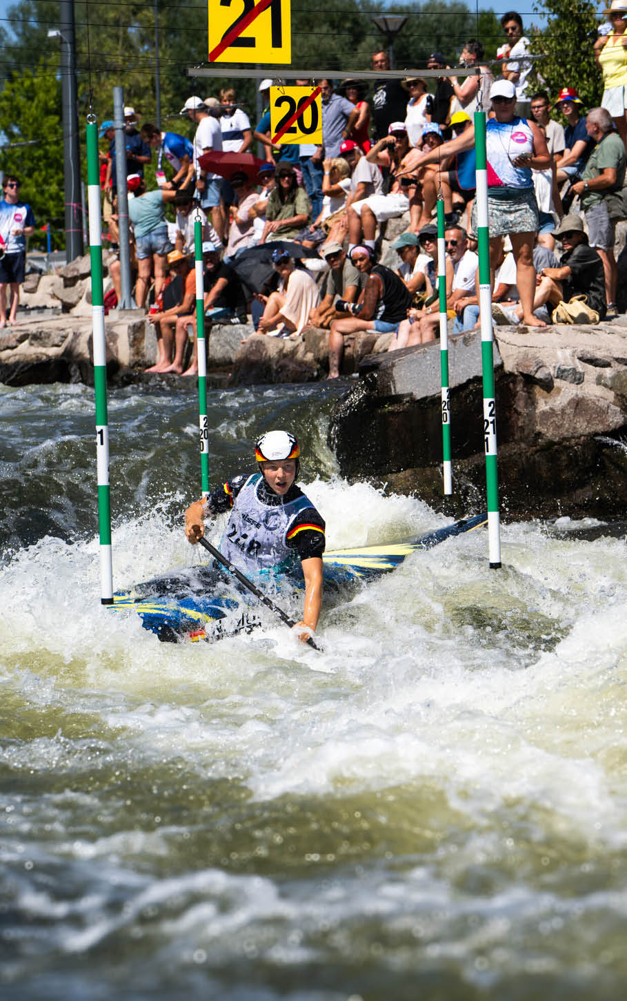 Eine Person in Sportkleidung und Helm f hrt mit einem Kajak durch einen Wildwasserparcours, w hrend zahlreiche Menschen am Ufer zuschauen. Beschreibung von KI generiert