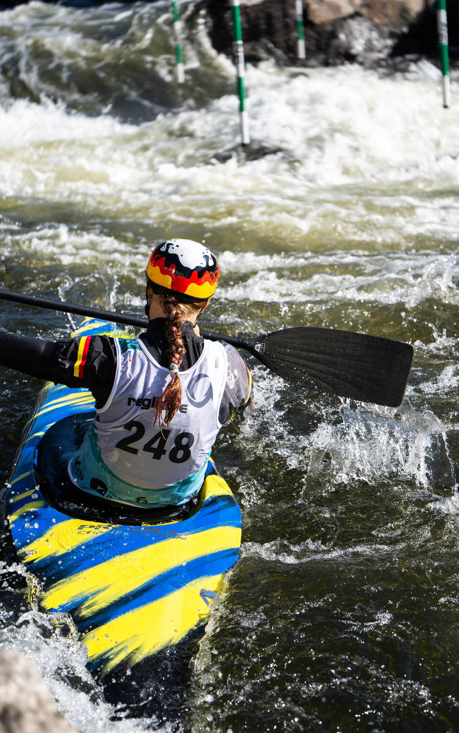 Eine Person mit Helm und Trikot paddelt in einem bunten Kajak durch wildes Wasser bei einem Kanuslalom-Wettbewerb. Beschreibung von KI generiert