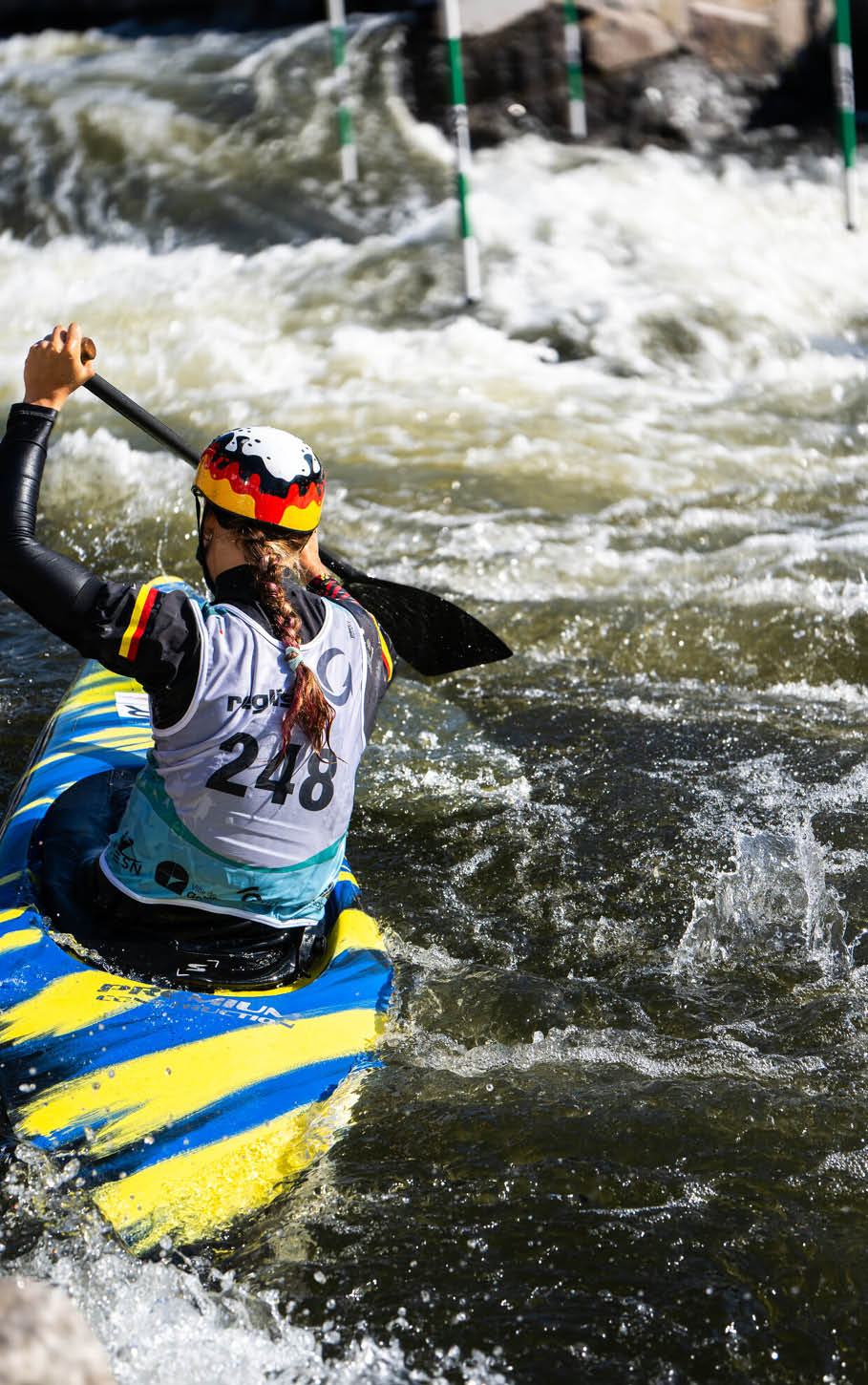 Eine Person mit Helm und Trikot paddelt in einem gelb-blauen Kajak durch wildes Wasser bei einem Kanuslalom-Wettbewerb. Beschreibung von KI generiert