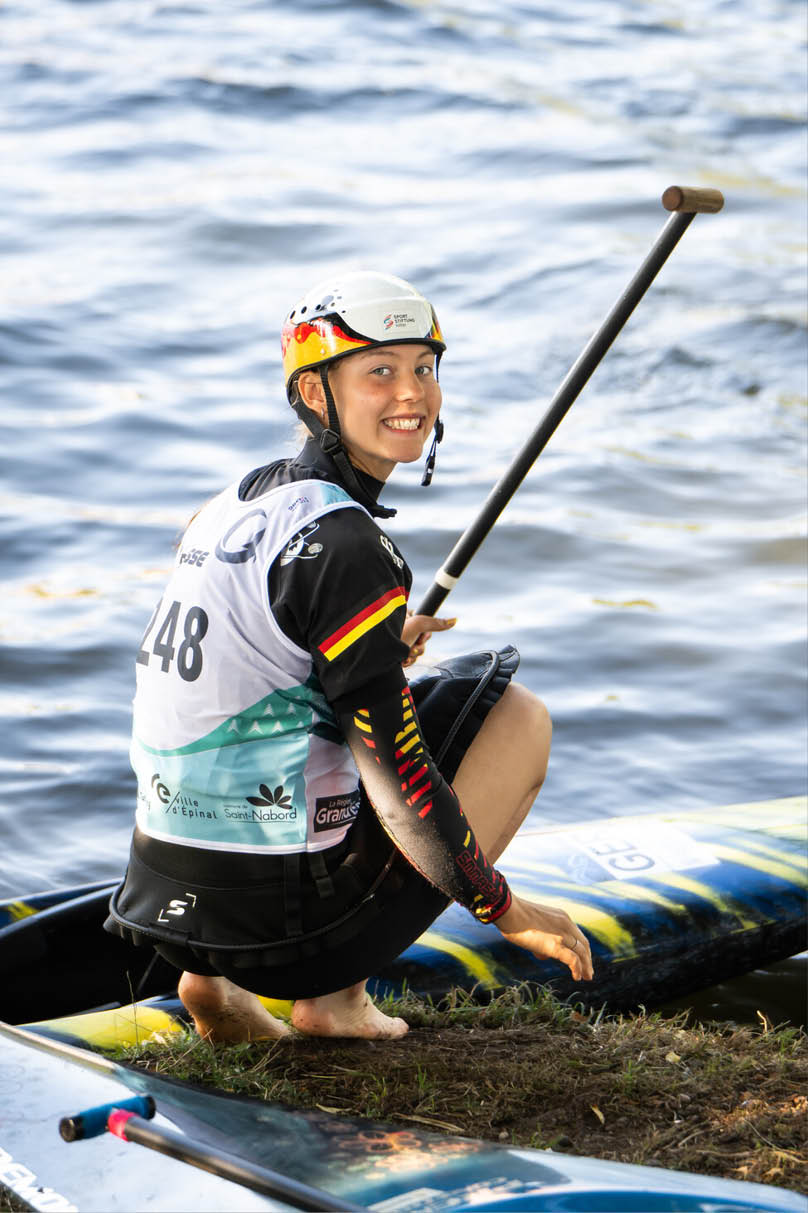 Eine Person mit Helm und Sportbekleidung sitzt auf einem Paddleboard auf einem Gew sser und h lt ein Paddel. Beschreibung von KI generiert