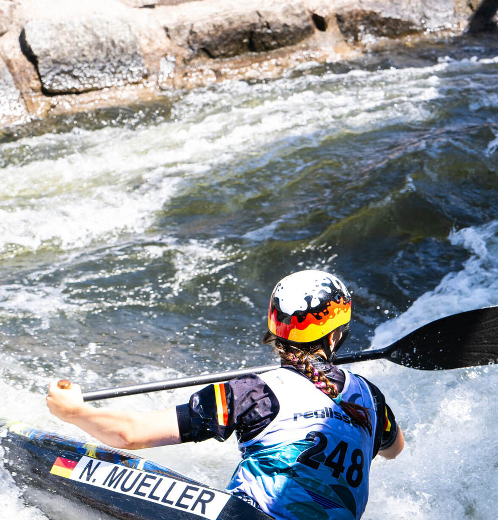 Eine Person mit Helm und geflochtenem Haar paddelt in einem Wildwasserfluss. Beschreibung von KI generiert