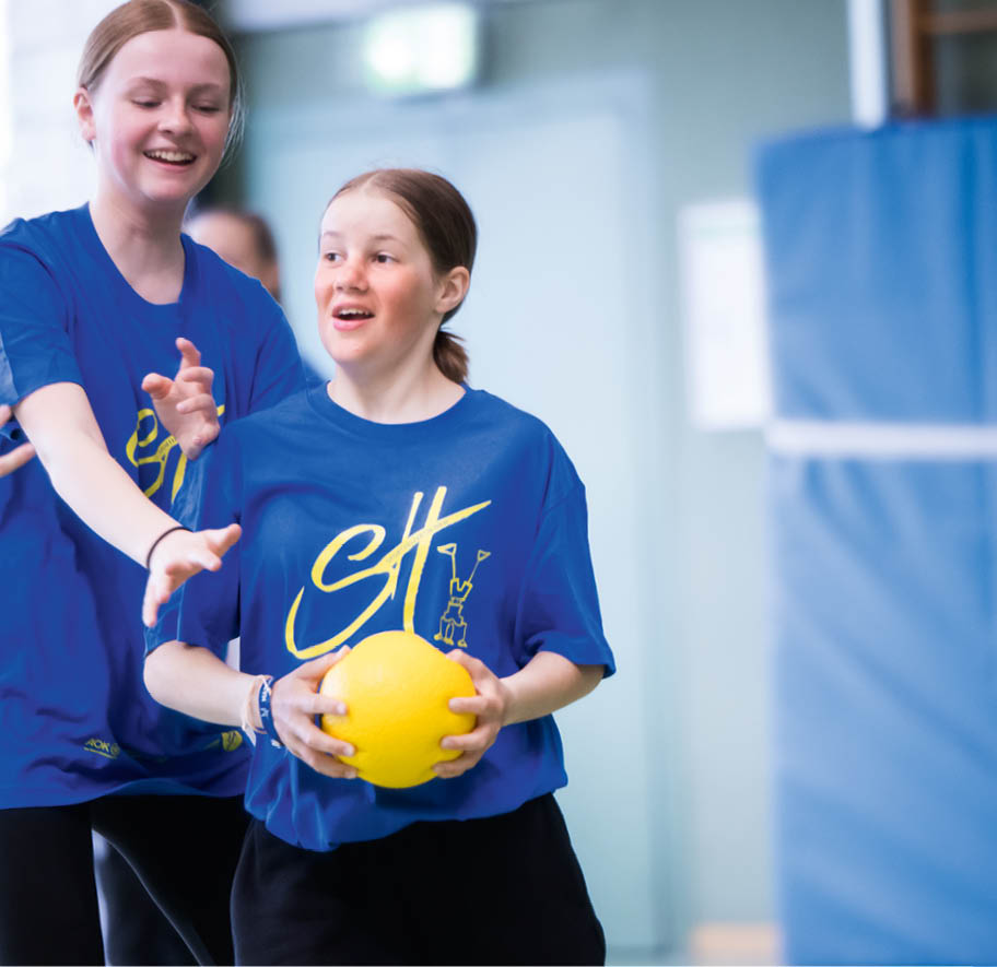 Zwei Personen in blauen T-Shirts spielen gemeinsam mit einem gelben Ball in einer hellen Sporthalle. Beschreibung von KI generiert