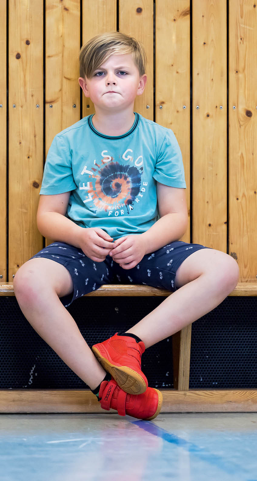 Ein Kind mit kurzem Haar sitzt in einem t rkisfarbenen T-Shirt und dunklen Shorts mit roten Sportschuhen vor einer Holzverkleidung und schaut ernst.