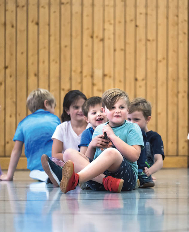 F nf Kinder sitzen in einer Reihe auf dem Boden eines Raums mit Holzwand, wobei das vorderste Kind ein Spielzeug h lt und alle entspannt wirken. Beschreibung von KI generiert