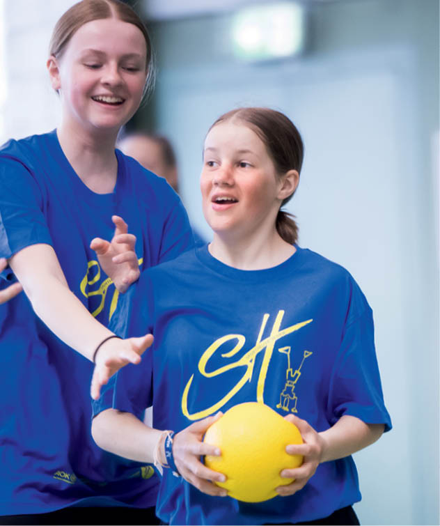 Zwei Personen in blauen T-Shirts spielen gemeinsam mit einem gelben Ball in einer hellen Sporthalle. Beschreibung von KI generiert