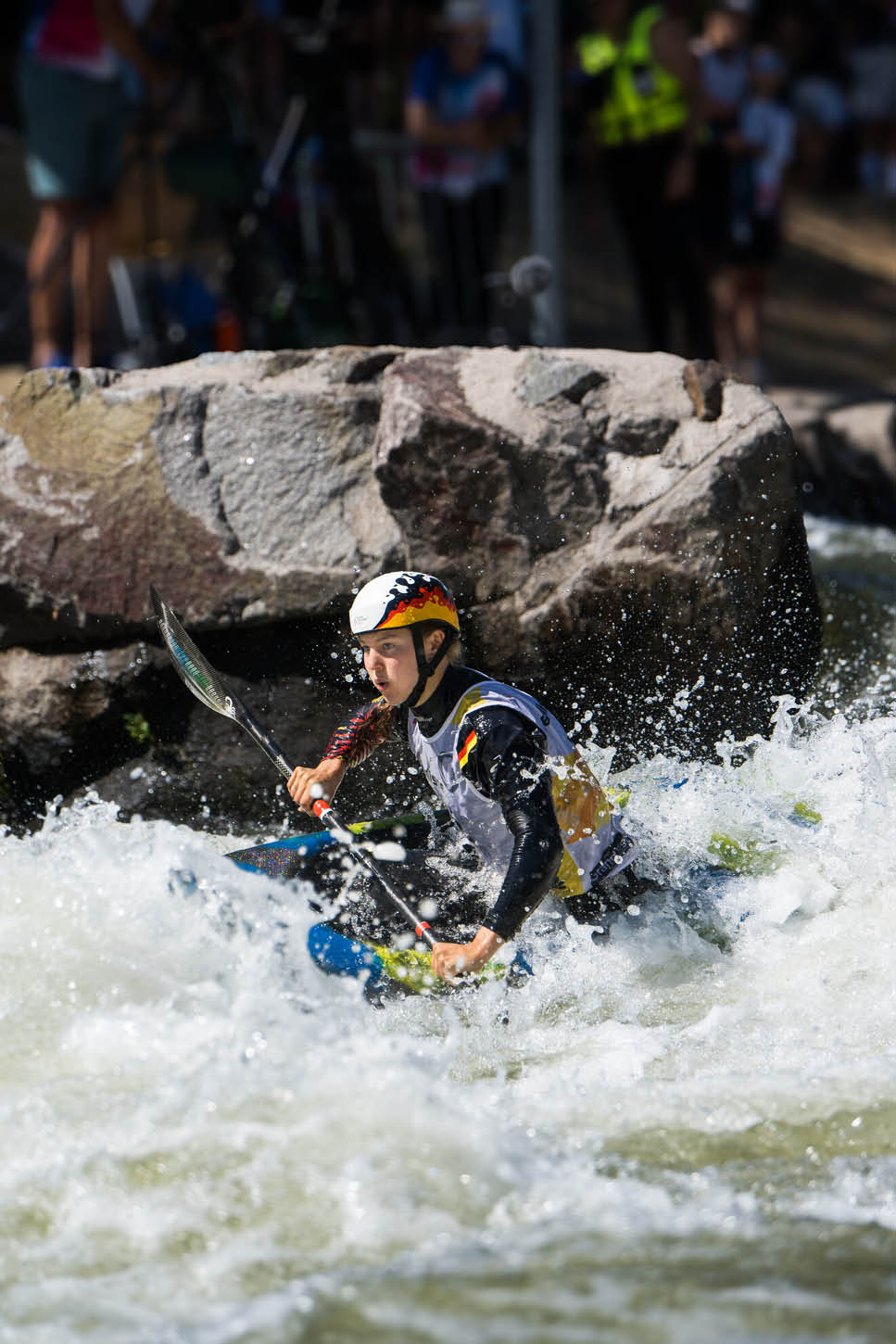 Eine Person mit Helm und Schwimmweste paddelt konzentriert in einem Kajak durch wildes, spritzendes Wasser vor einem gro en Felsen. Beschreibung von KI generiert