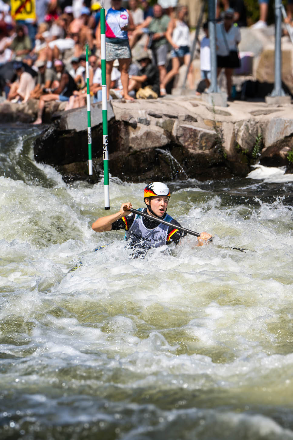 Eine Person tr gt einen Helm und Paddel und f hrt in einem Wildwasserfluss durch ein Tor bei einem Kanuslalom-Wettbewerb vor einer Zuschauergruppe. Beschreibung von KI generiert