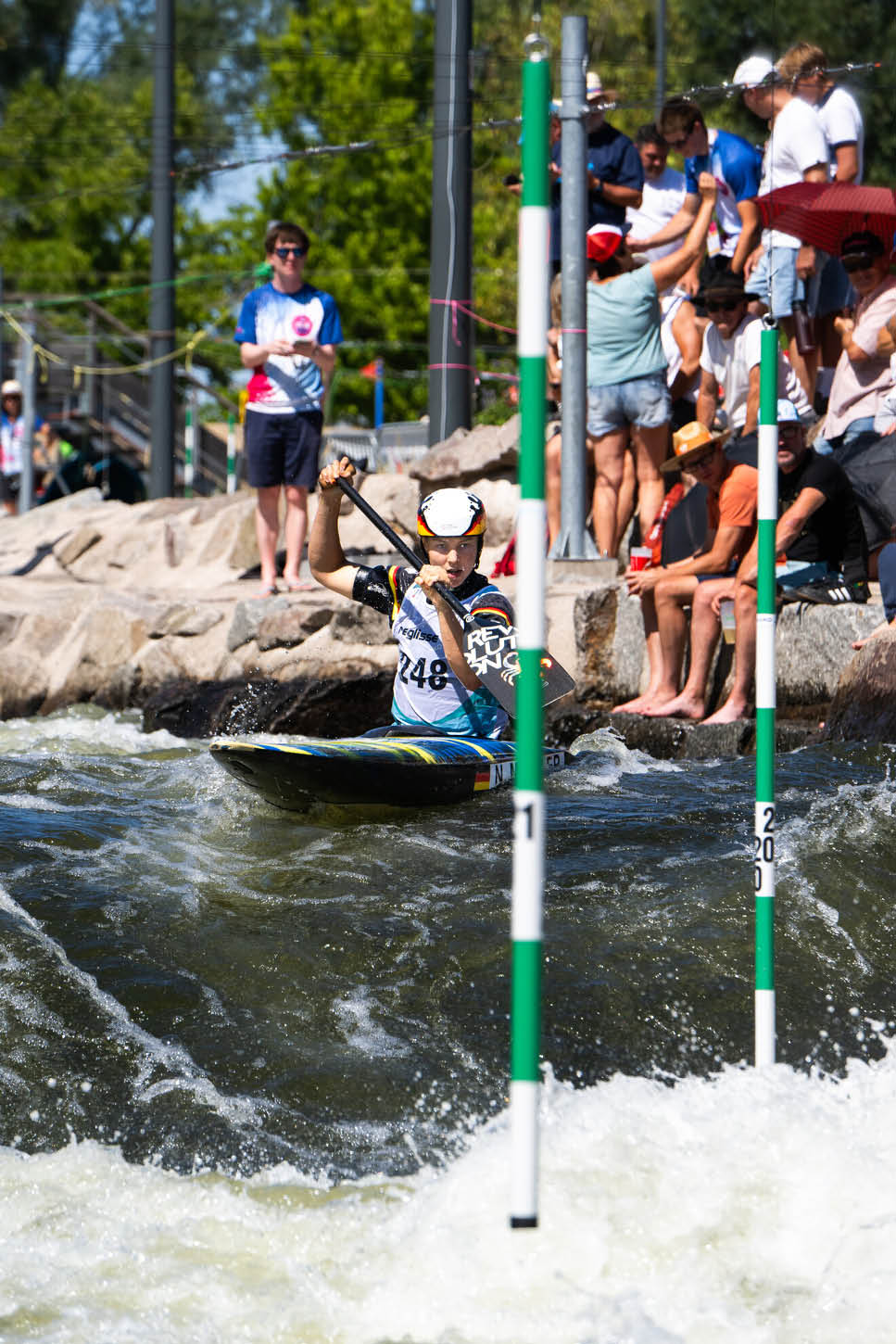 Eine Person mit Helm und Sportbekleidung paddelt in einem Kajak durch Wildwasser, w hrend mehrere Menschen am Ufer zuschauen. Beschreibung von KI generiert