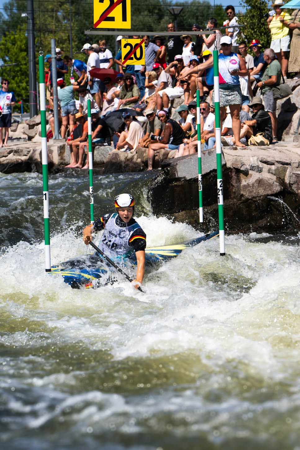 Eine Person in Sportkleidung und Helm f hrt mit einem Kajak durch einen Wildwasserparcours, w hrend zahlreiche Menschen am Ufer zuschauen. Beschreibung von KI generiert